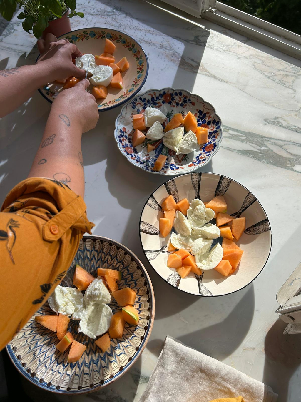 Hands plating melon and mozzarella into patterned bowls on a sunlit marble counter