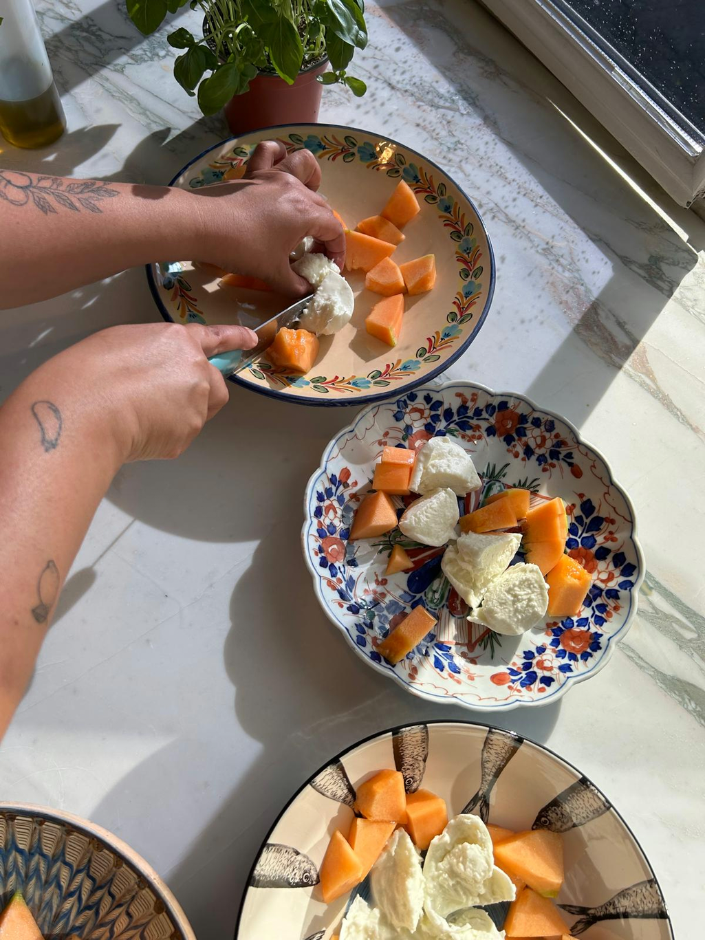 Hands plating melon and mozzarella onto patterned plates in sunlight